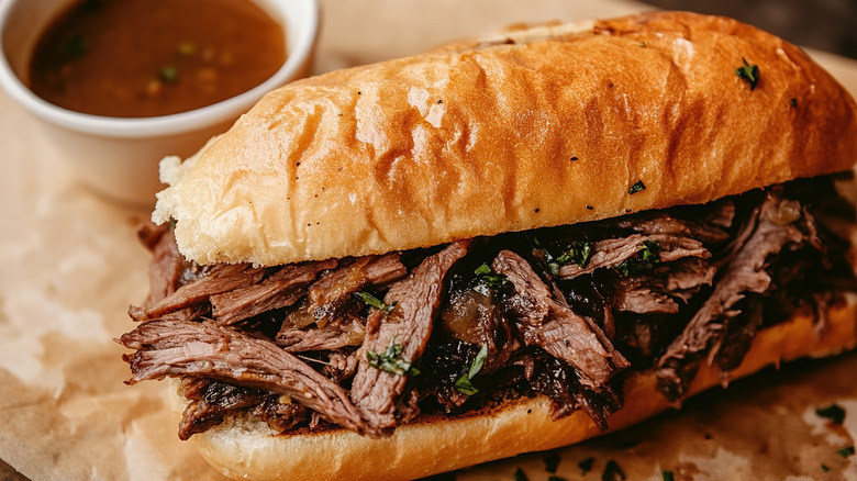 Sliced pieces of beef on a long roll with a small bowl of brown dipping juices in the background