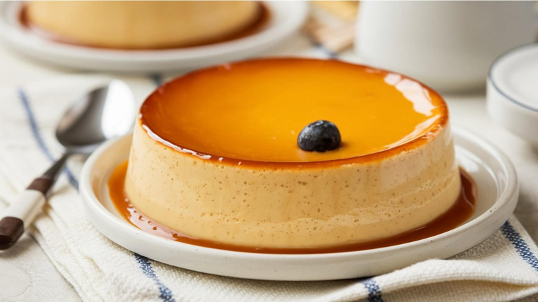 A flan with one blueberry on top sits on a white plate