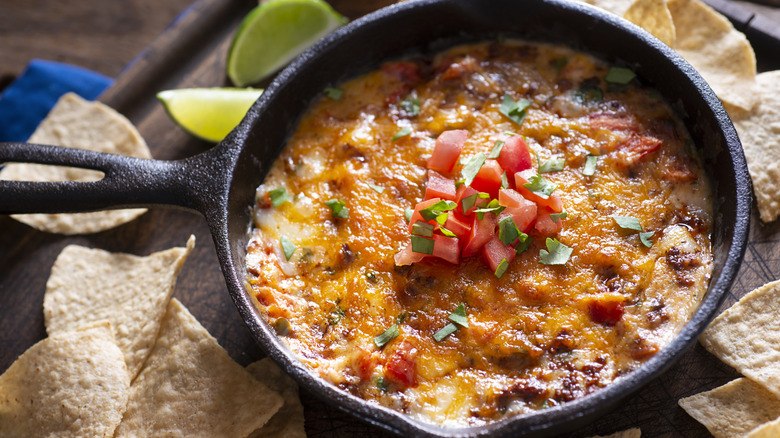 A skillet of queso with chorizo and diced tomatoes sits on a wood table surrounded by tortilla chips