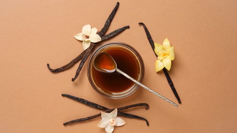 A bowl of vanilla extract with a spoon and whole vanilla pods with flowers on the side