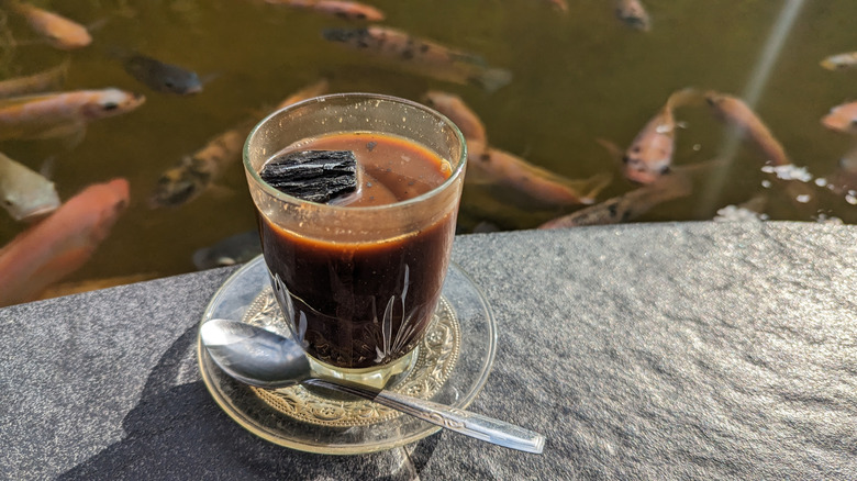 Clear mug on small saucer with spoon atop a concrete table, filled with kopi joss. Black charcoal floating on top with koi fish swimming in the background