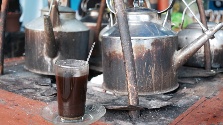 A clear cup of coffee with charcoal, set in front of worn metal coffee pots on an old stove