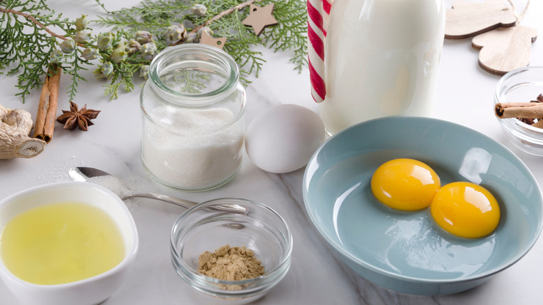 Eggnog ingredients, raw egg, sugar, spice, and milk in jars and bowls on a white table