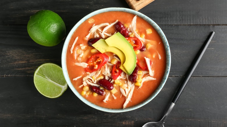 Bowl of Mexican soup with shredded chicken avocado and chilis with lime on dark wood background