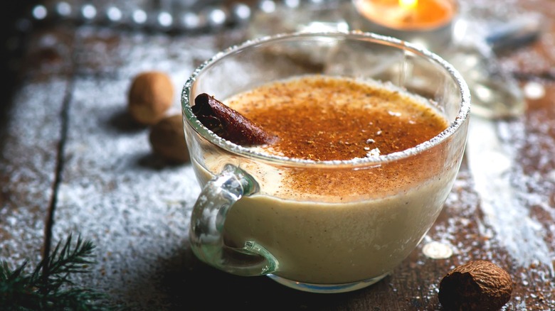 A glass of eggnog held by a see-through teacup with cinnamon and a cinnamon stick inside