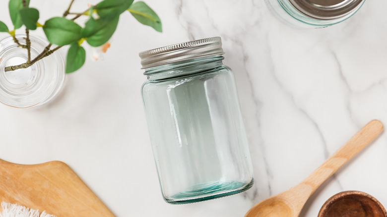 An empty mason jar is lying flat on a white marble table, and around it are a plant, a wooden baking spoon, and a wooden cutting board.