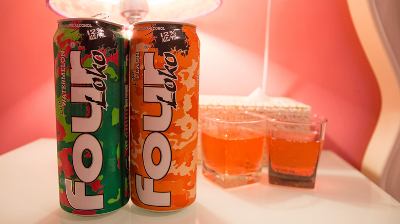 Two cans of Four Loko sit on a nightstand next to two shot glasses full of orange liquid