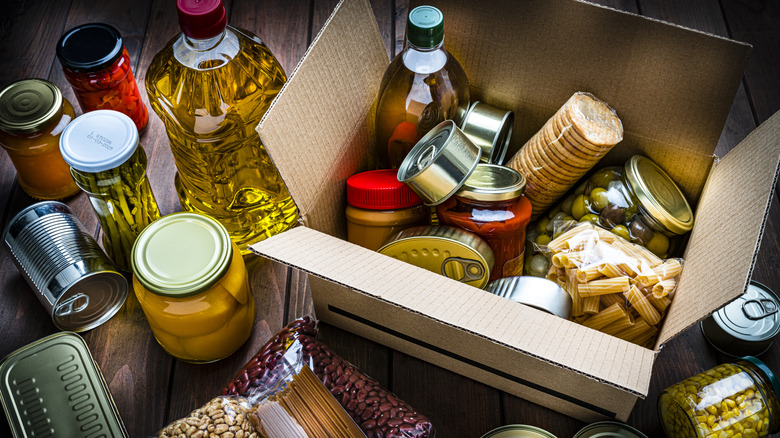 Various shelf-stable foods like crackers, produce, and metal tins in a box and on wooden floor