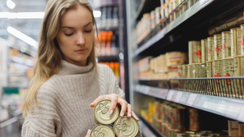 A woman holding three cans of food stands in front of a canned goods shelf in a store