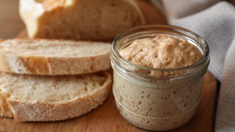 sourdough bread starter dough in a jar sits next to a loaf of sliced sourdough