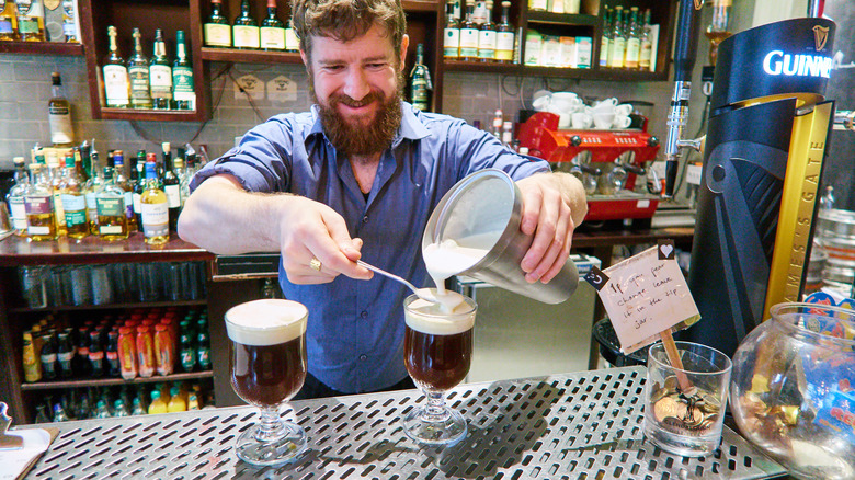 A smiling bartender pours whipped cream into a glass filled with Irish coffee