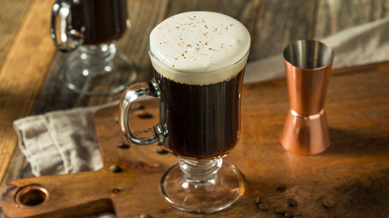 An Irish coffee topped with whipped cream sits on a wooden table next to a copper shot glass