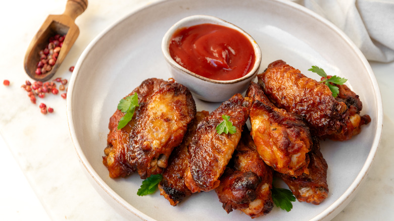 A plate of crispy chicken wings next to a bowl of sauce and some peppercorns