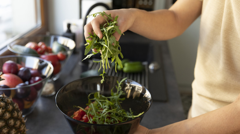 A person tosses a handful of arugula into a black salad bowl with chopped tomatoes