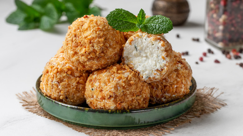 Pile of cheese balls on a green, ceramic plate. One cheese ball has been cut open, showing cream cheese interior. Topped with garnish and all on a placemat and white table with seasonings, and green plant blurred in the background.