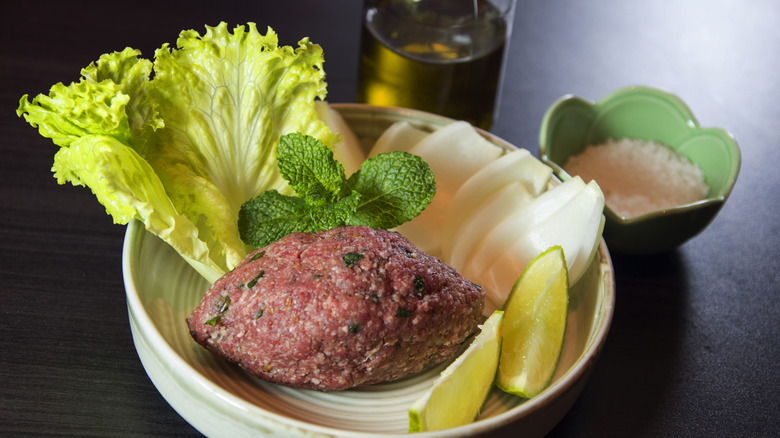 Lebanese steak tartare garnished with mint, endive, onion, lime wedges, near a bowl of salt and olive oil