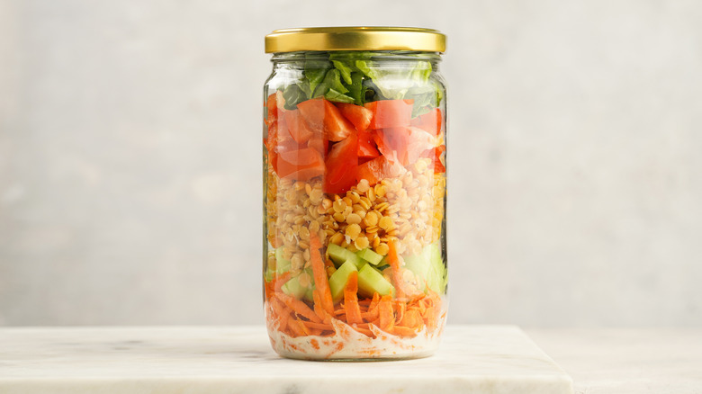 A mason jar with layered salad ingredients including salad dressing, carrots, cucumbers, yellow lentils, tomatoes, and lettuce