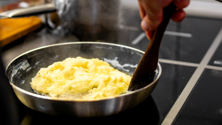 A cook stirs scrambled eggs with a wooden spatula on an electric stove