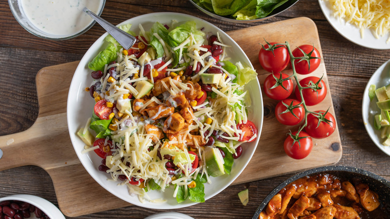 A Tex-Mex salad in a white bowl sits on a cutting board next to cherry tomatoes, a dish of dressing, and other salad ingredients