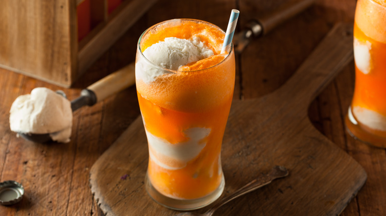 Creamsicle frozen cocktail in tall glass with ice cream shown with orange soda and straw at the top. Ice cream scoop with vanilla ice cream on the left side, and spoon on the right. All on a wooden table.