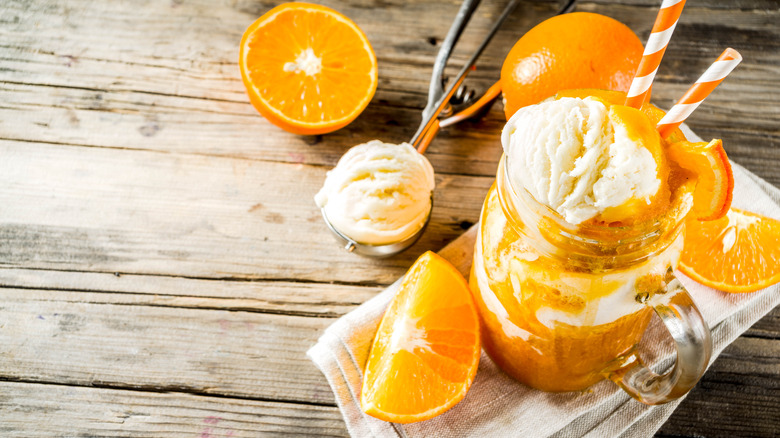 Creamsicle frozen cocktail in a mason jar with two striped straws. Orange slices and whole orange surrounding glass, with ice cream scooper with vanilla ice cream on the upper left. All on a light, wooden table.