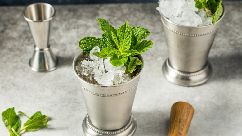 Two mint juleps in silver cups with crushed ice and mint leaves piled on top on a light gray surfaces with a silver jigger, mint leaves, and a wooden muddler on the side