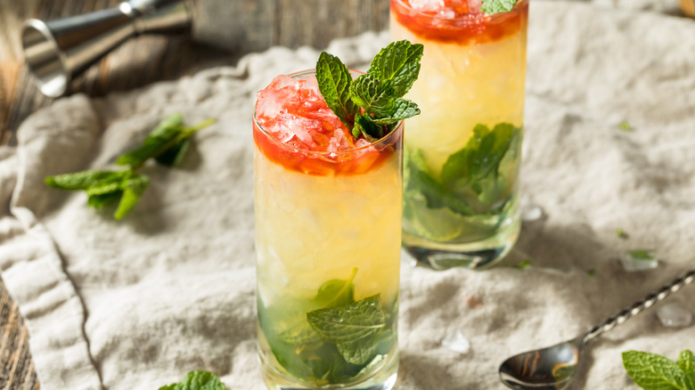 Two light yellow drinks with red on top and lots of crushed ice with mint leaves on the bottom of the glass and sticking out on a cream towel with a metal jigger on its side in the background and a long metal cocktail spoon and mint leaves in the foreground