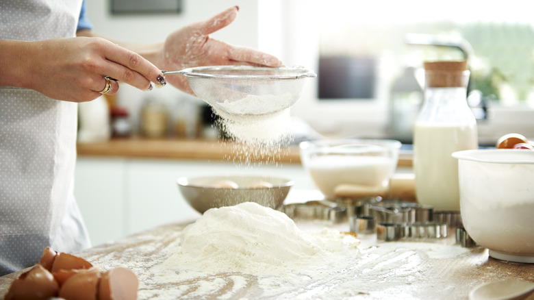 Hands sift flour through a mesh strainer onto a kitchen countertop