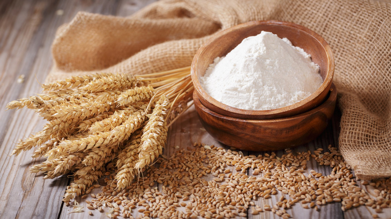 A wooden bowl of flour sits on a piece of burlap near scattered whole grains and stalks of wheat