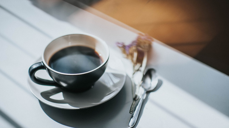 A small cup of black coffee sitting on a white saucer next to a spoon and a dried flower