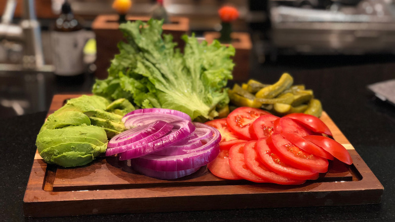 Various burger toppings sit on a wooden cutting board, including lettuce, pickles, tomato, red onion, and avocado