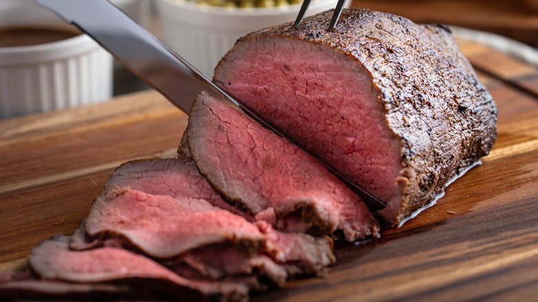 Roast beef with a knife cutting into it on a wooden cutting board