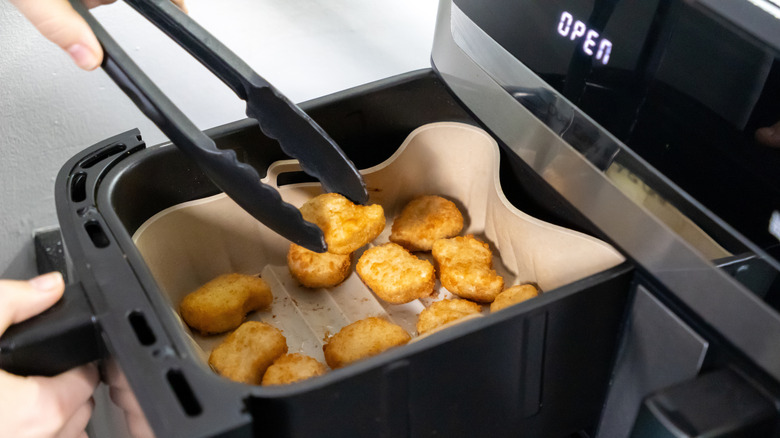 A hand uses tongs to turn chicken nuggets in an air fryer basket