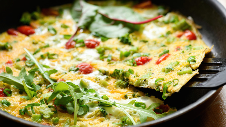 Vegetable frittata with tomatoes and scallions, with herbs on top in a black pan with one triangular piece being lifted out by a black spatula