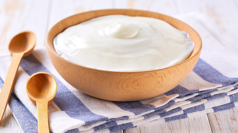 A bowl of plain Greek yogurt sits on a folded blue and white striped cloth with wooden spoons on the side.