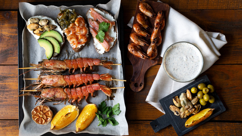 A seacuterie board with grilled langoustines, smoked mussels, lox, canned fish, prawns, and shrimp all beside a mix of fresh and roasted fruit, garlic, herbs, olives, a dipping sauce, and slices of toast