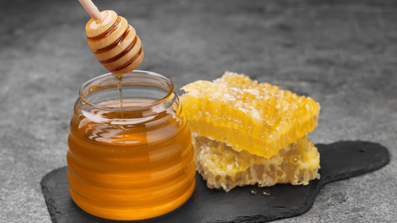 A jar of honey with honey dripping from the honey dipper on a slab with honey comb