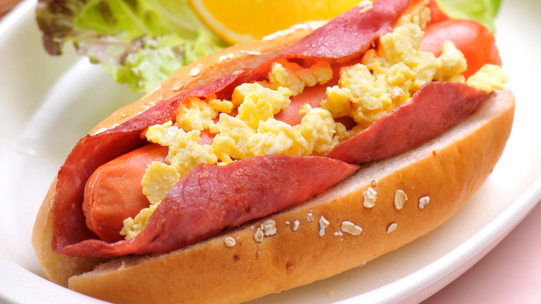 Hot dog on cooked ham on a bun with scrambled eggs on top resting on a white plate