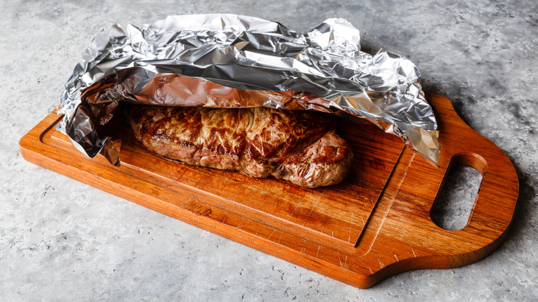 Steak covered in foil on a wooden cutting board