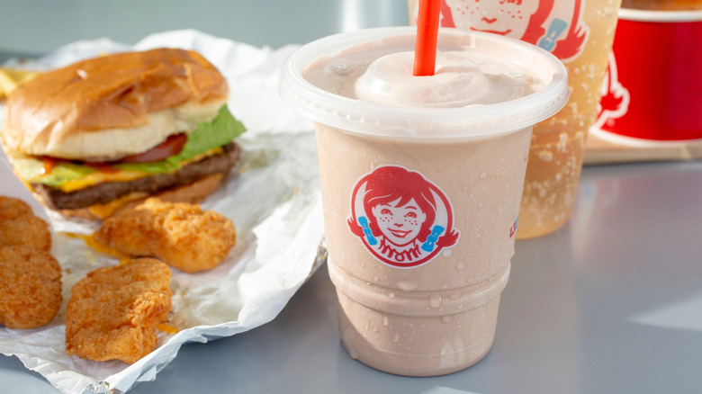 Wendy's burger and three chicken nuggets on an open wrapper with a cream-colored Frosty with a straw and the Wendy's logo on front next to it