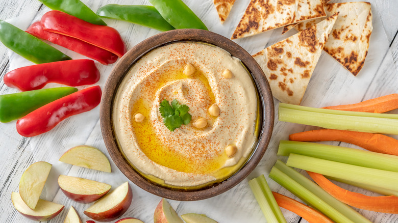 A bowl of hummus is surrounded by a selection of fruits, vegetables, and pita bread