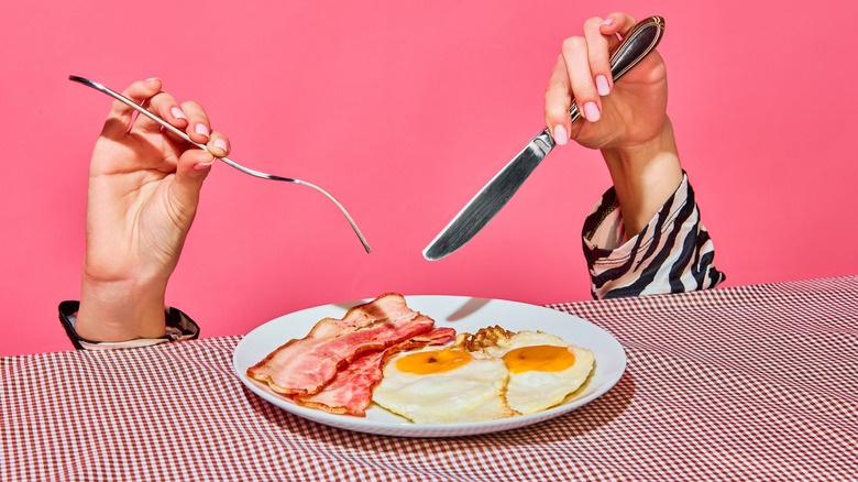 bacon and egg breakfast plate with hands holding knife and fork