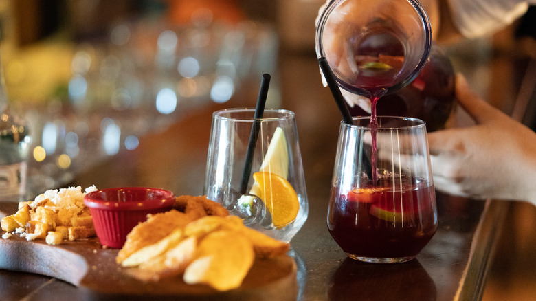 Person pouring cocktail into glass next to wooden board of appetizers