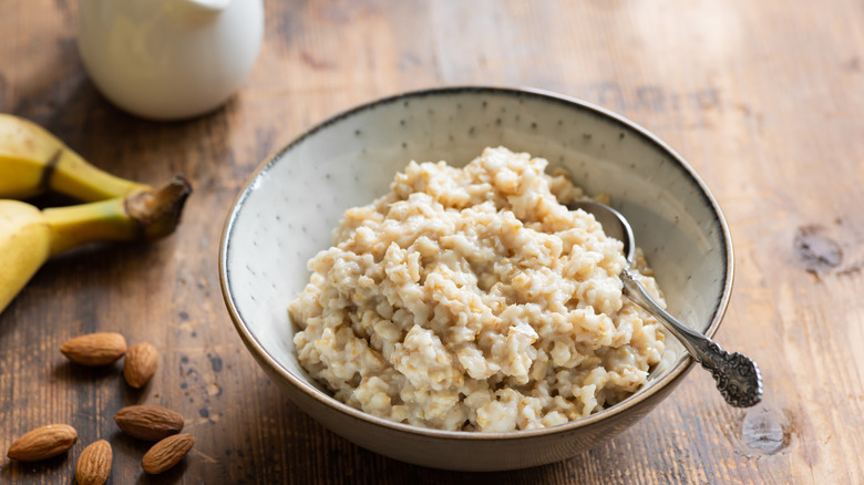 A bowl of oatmeal placed at a table next to bananas, almonds, and milk