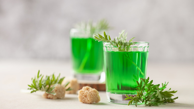 Two glasses of green absinthe with sugar cubes, and sprigs surrounding, on a texturized, light background.