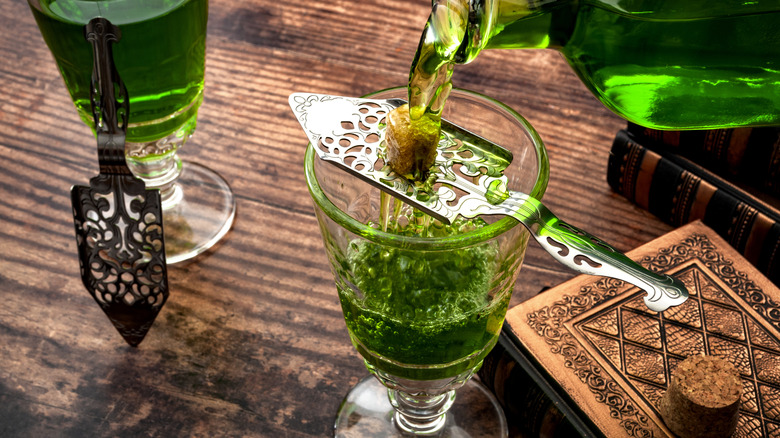 Bottle of absinthe being poured over a sugar cube on a metal spoon into a glass, with a book and cork in the bottom right corner. All on a wooden table.