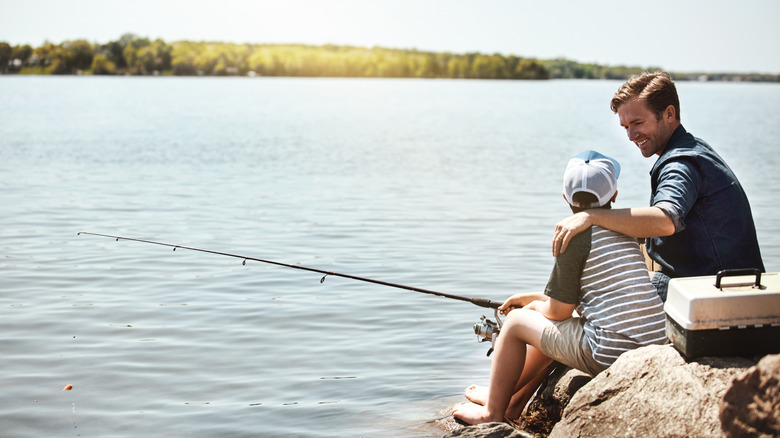 Happy father, fishing and lesson with son on rock with rod by lake, ocean or beach together in nature. Dad with kid, young child or little boy for bonding, teaching or tips to catch by sea water