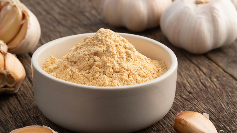 Close up of garlic powder in a small white bowl with cloves of garlic surrounding.