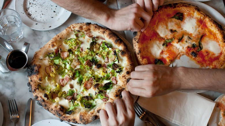 People grabbing pizza slices from two pizzas on a table