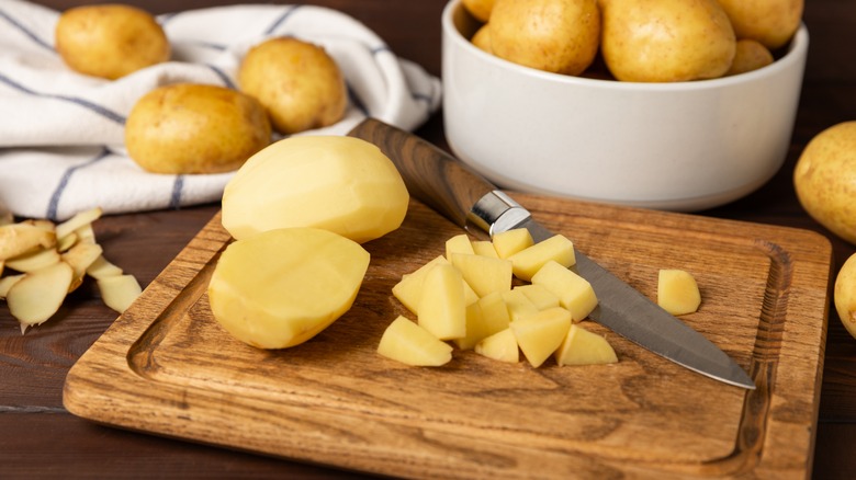 Raw potatoes, some peeled and chopped up, with a knife on a wooden cutting board.